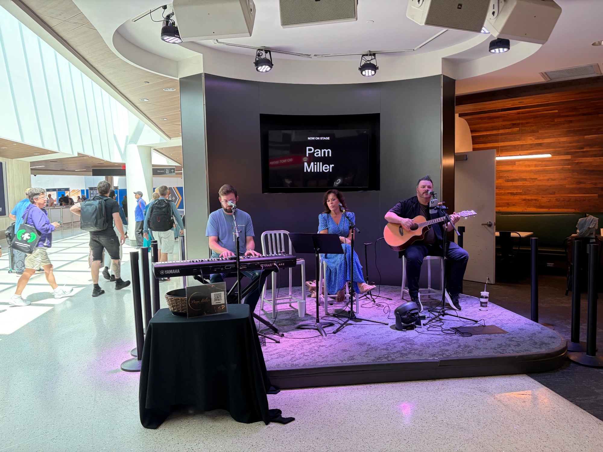 Four musicians playing on a small airport stage beneath a screen reading Pam Miller