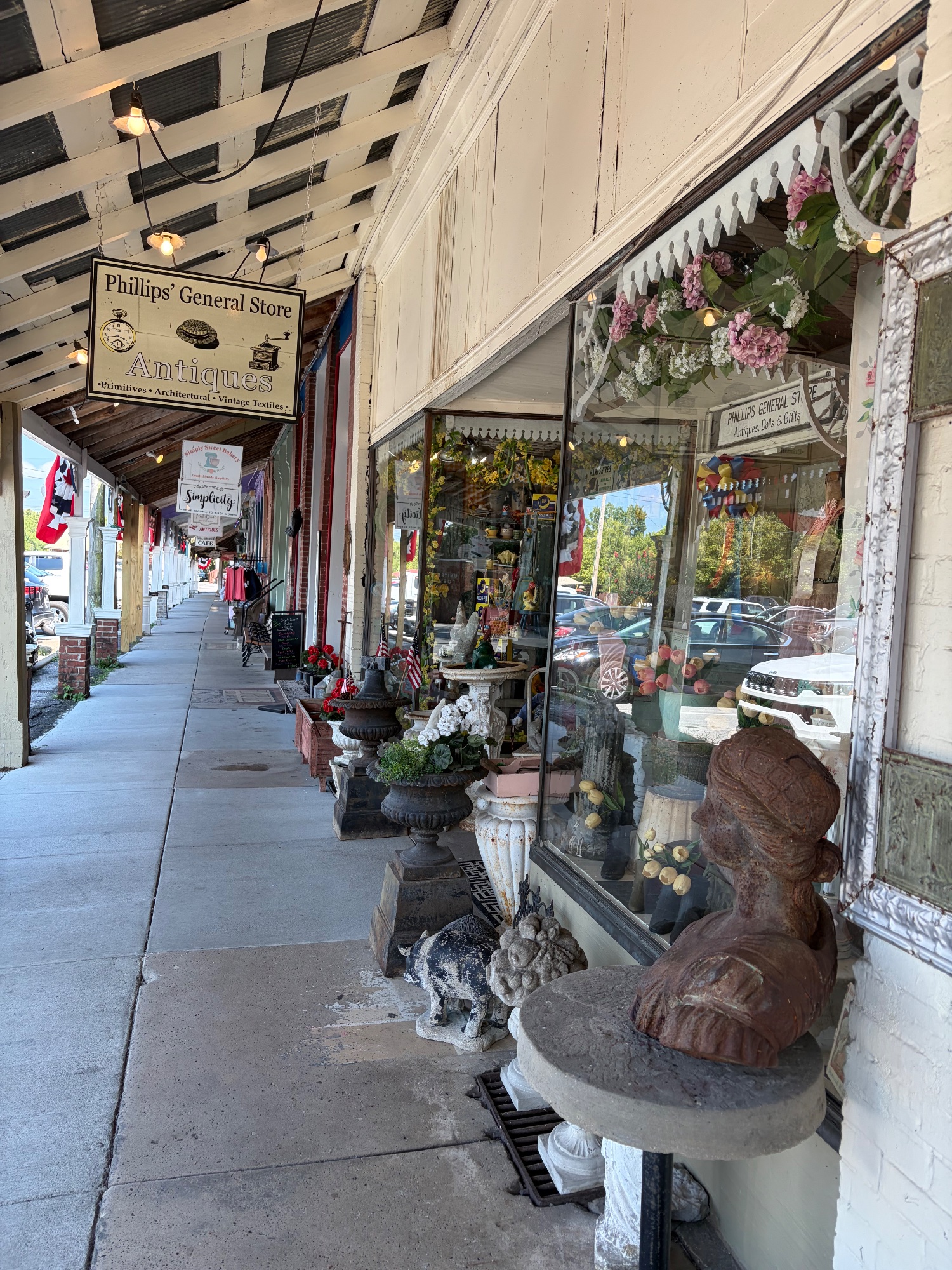 Covered wooden sidewalk past an antiques shop window filled with ornaments and flowers