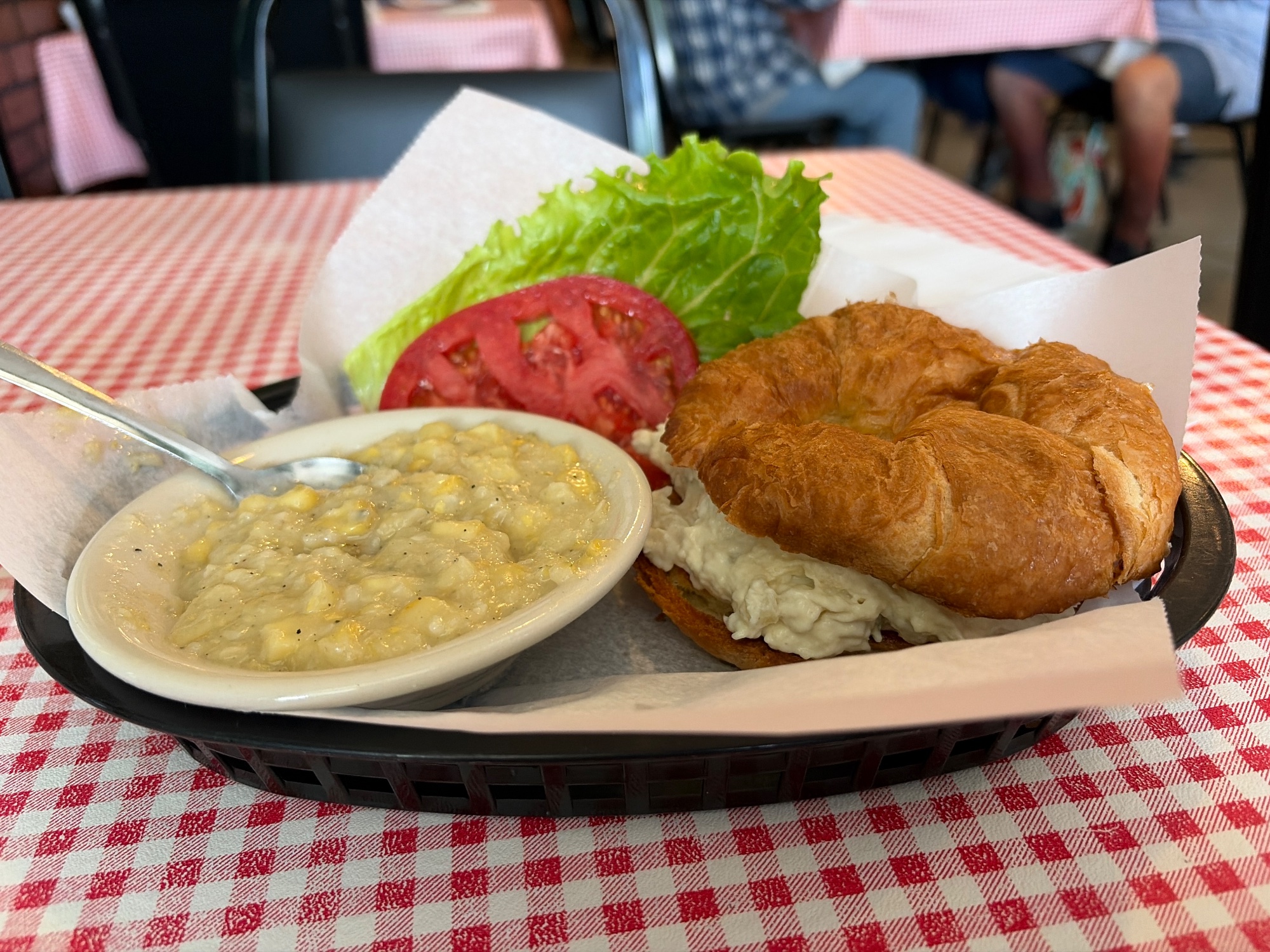 Basket meal of a biscuit with lettuce and tomato beside a bowl of corn chowder on a checked cloth