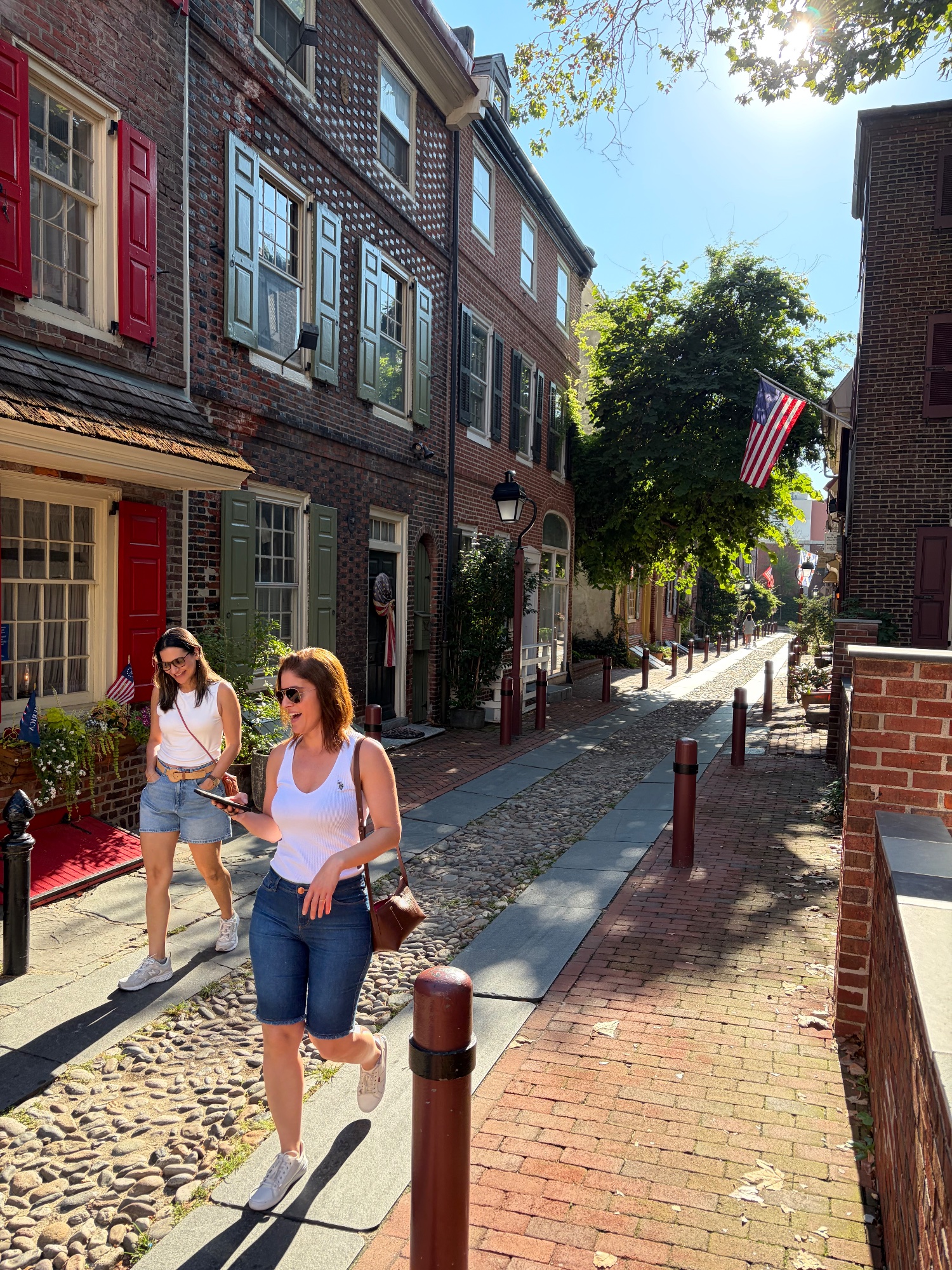 Two women walking down a narrow cobbled alley of brick houses with American flags overhead