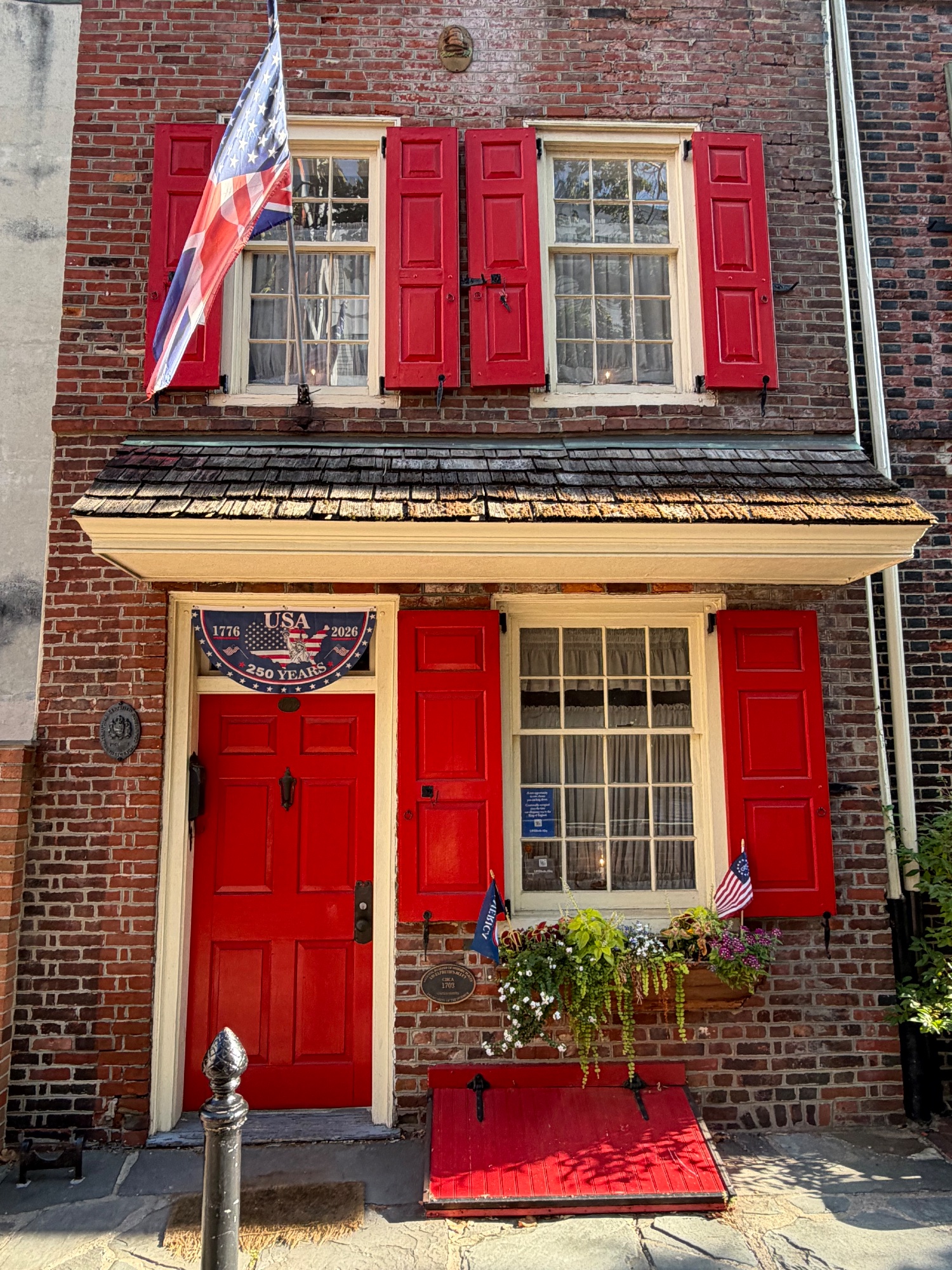 Red front door and shutters beneath a shingled pent roof with a 1776 to 2026 plaque beside it